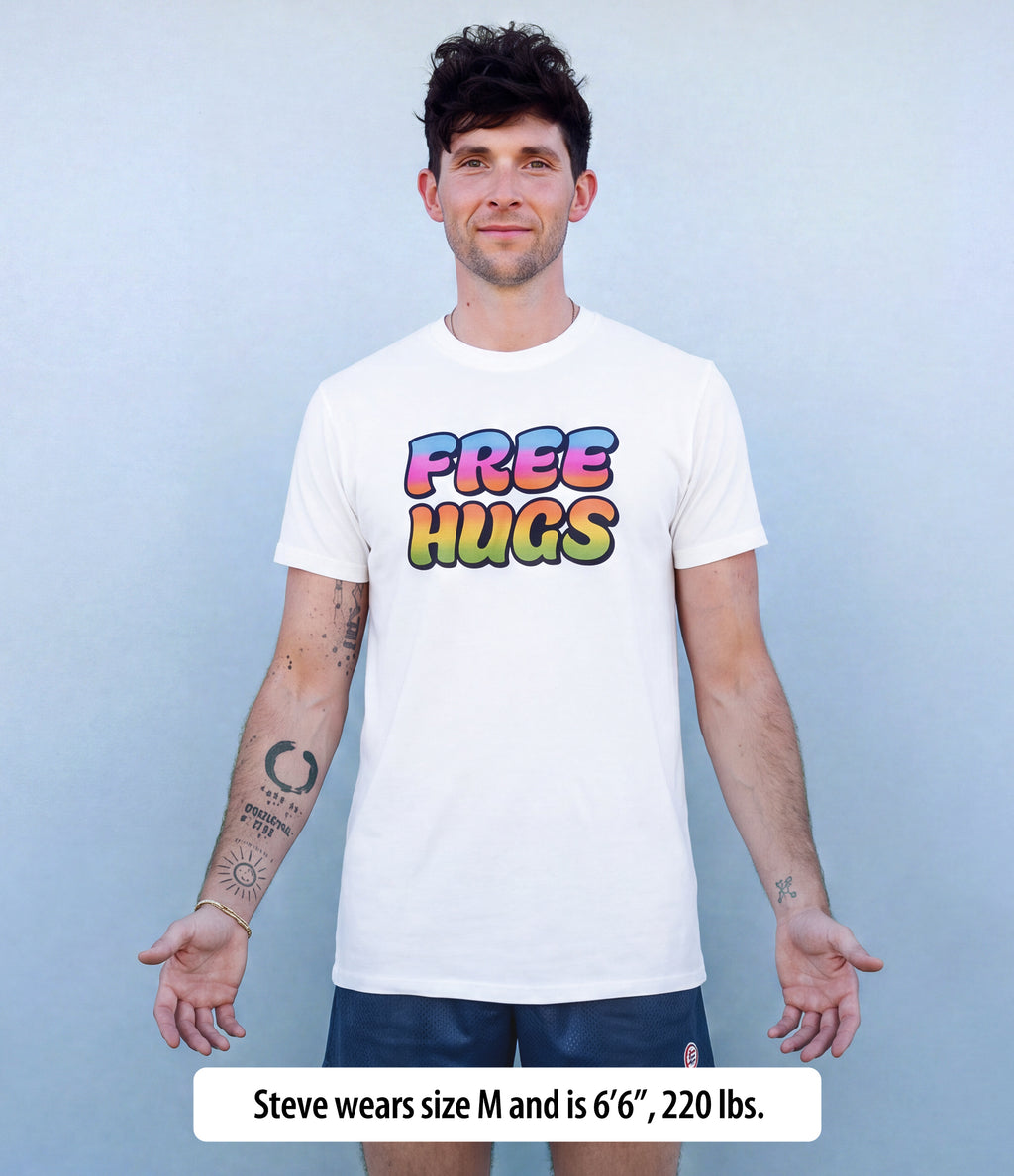 A man wearing a white crewneck t-shirt with a colorful 'FREE HUGS' graphic on the chest, standing against a light blue-gray background. A text overlay at the bottom reads 'Steve wears size M and is 6'6", 220 lbs.