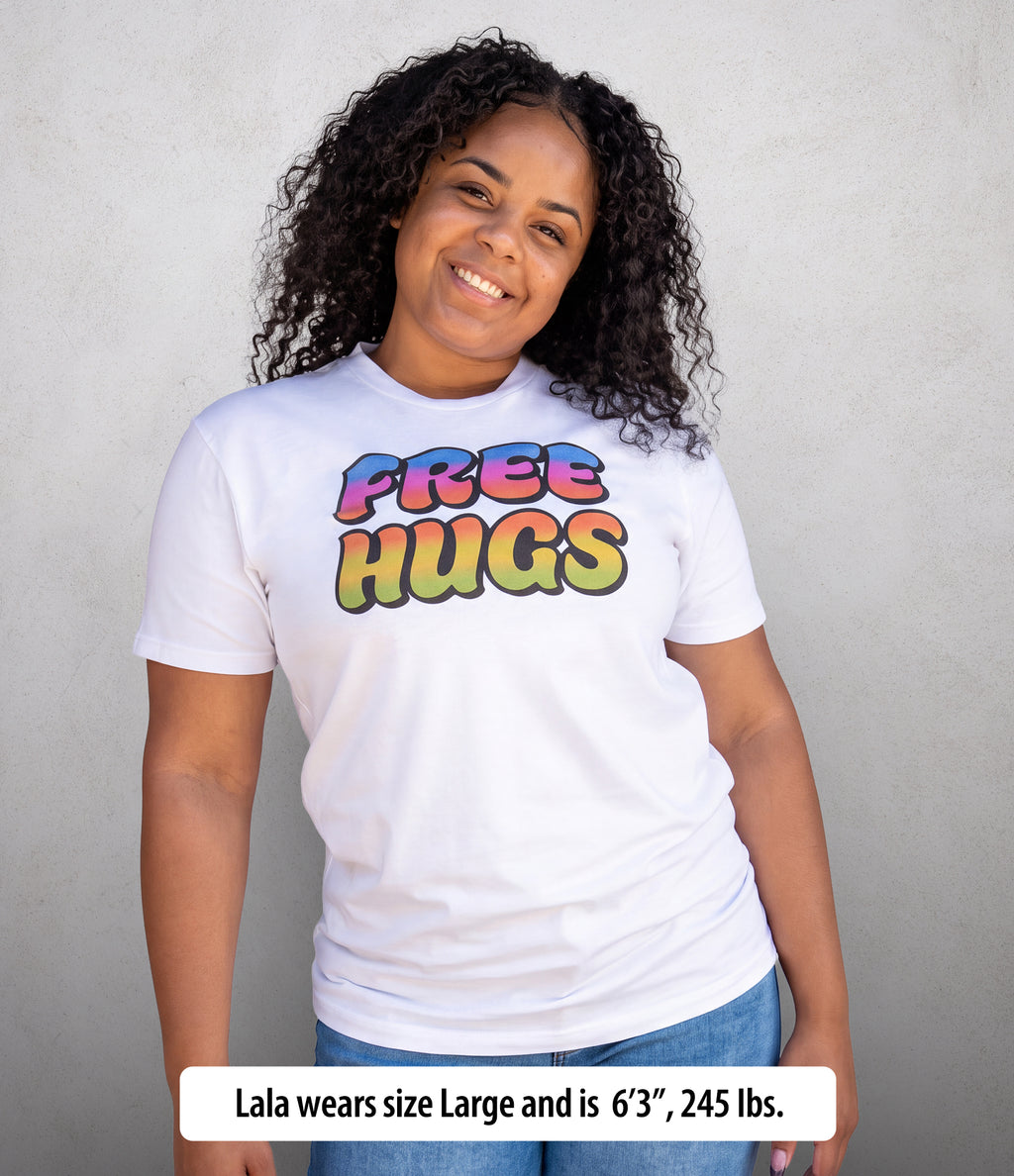 A smiling woman with curly dark hair wears a white short-sleeve T-shirt with a colorful 'FREE HUGS' graphic on the chest, paired with blue jeans, standing against a light gray textured wall. A text banner at the bottom reads, 'Lala wears size Large and is 6'3", 245 lbs.