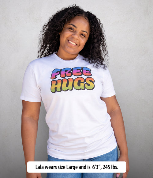 A smiling woman with curly dark hair wears a white short-sleeve T-shirt with a colorful 'FREE HUGS' graphic on the chest, paired with blue jeans, standing against a light gray textured wall. A text banner at the bottom reads, 'Lala wears size Large and is 6'3", 245 lbs.