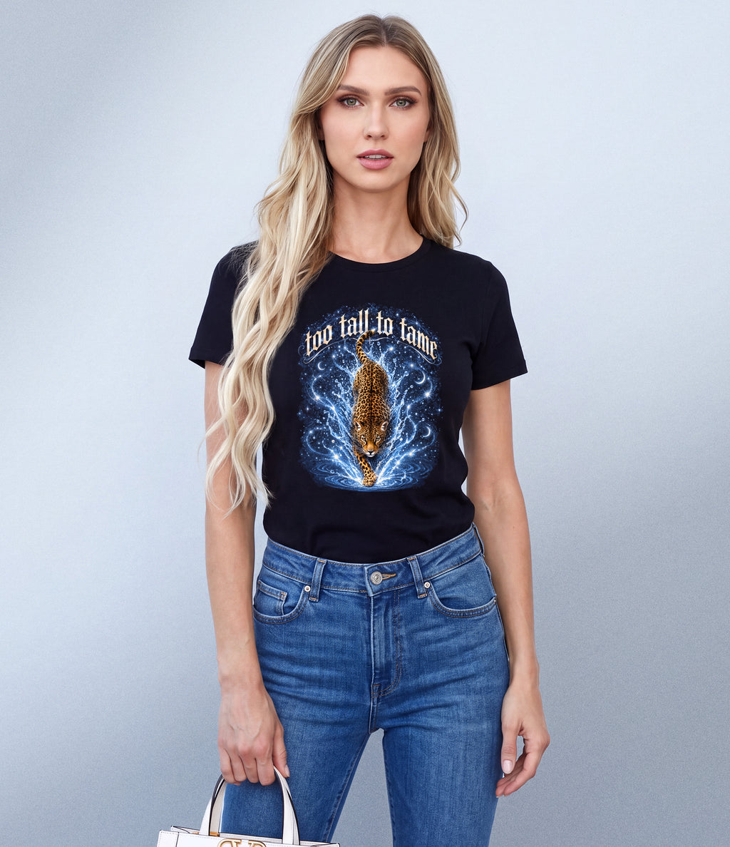 A blonde woman modeling a black graphic t-shirt with a blue and gold leopard print and the text 'too tall to tame', paired with blue jeans, against a light gray studio background.